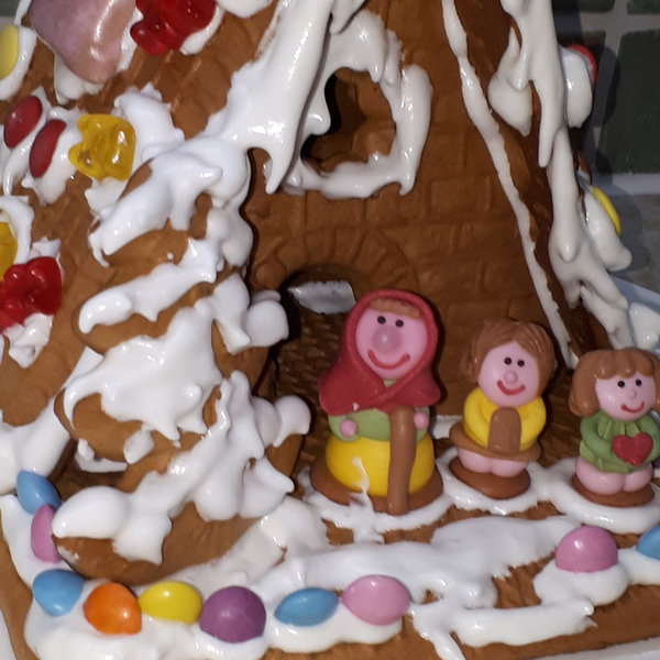 Pfefferkuchenhaus mit Zuckerguss, Samarties und Gummibärchen dekoriert. Vor dem Knusperhaus steht die Hexe mit Hänsel und Gretel aus Zuckerguss
