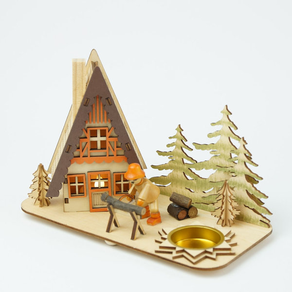 Teelichthalter mit einem Räucherhäuschen aus Holz im Fachwerkstil mit einer Holzfigur, welche sich über einen Sägebock beugt und Holz sägt. Im Hintergrund ist ein Holzstapel und 5 Tannen